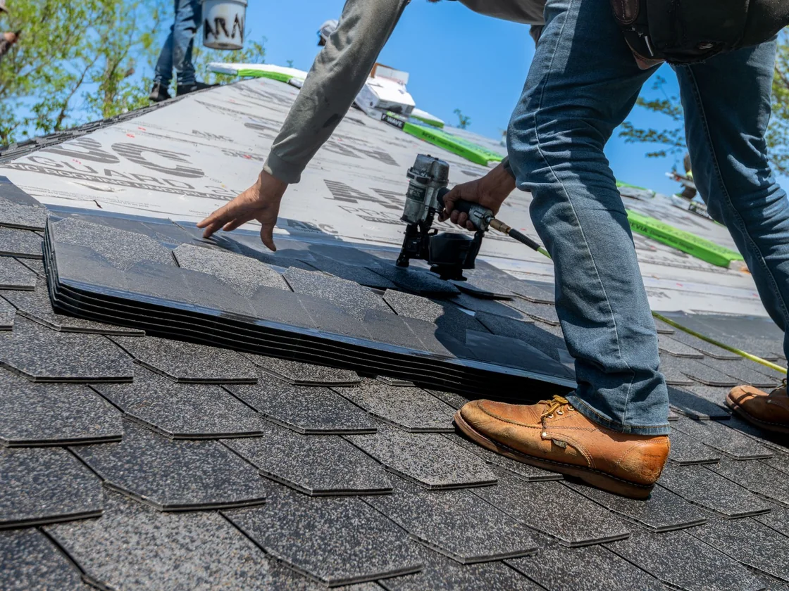 A roofer nailing down a fresh course of asphalt shingles over new underlayment on an ordinary house roof