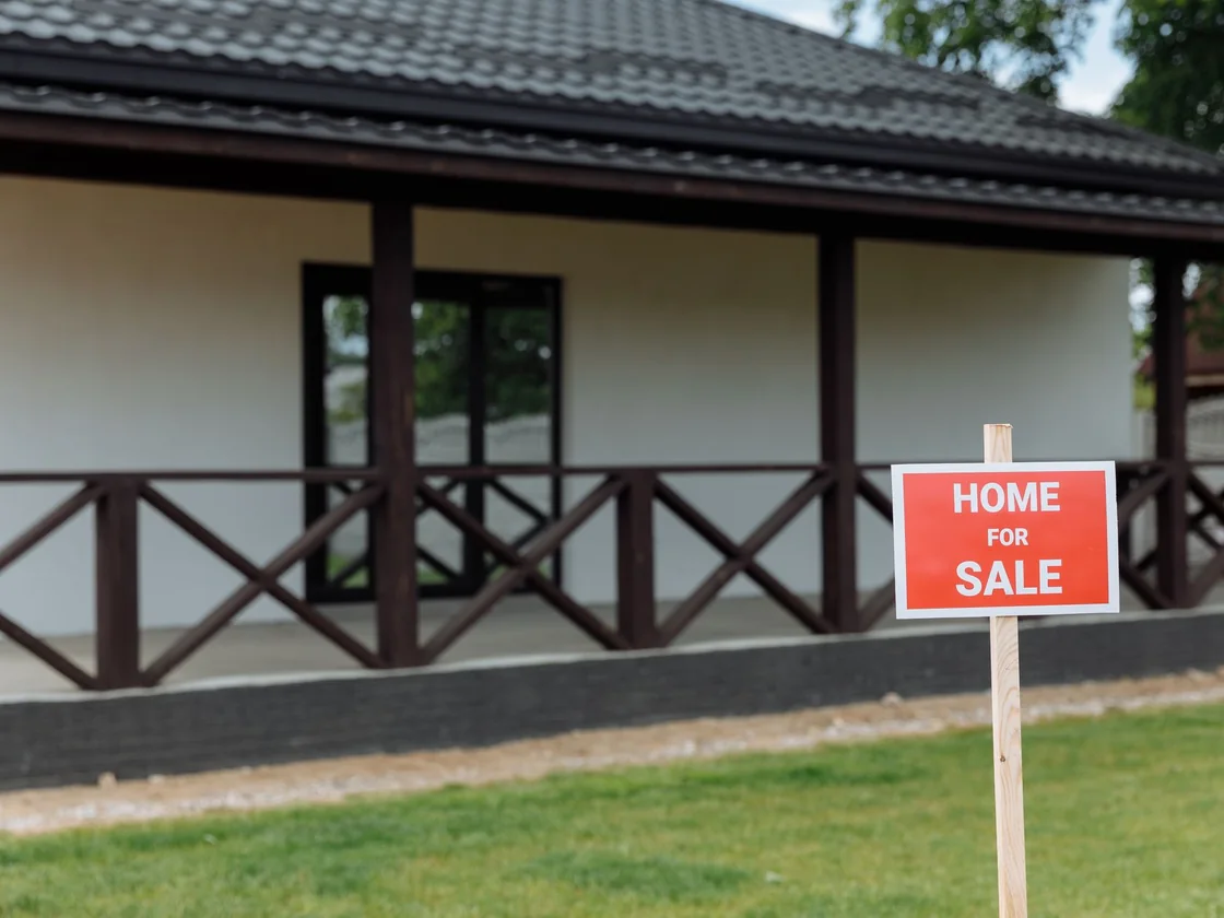 A red home for sale sign on a wooden stake in the lawn of a plain single story house with a covered porch