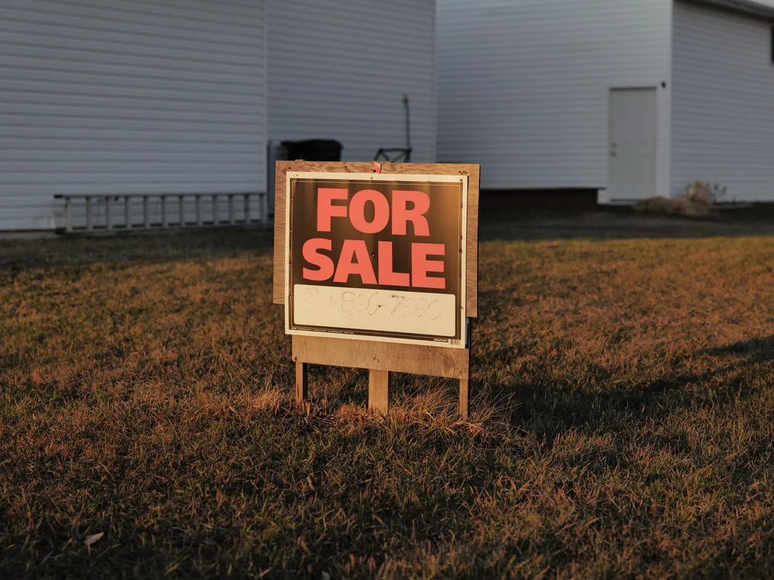 A homemade for sale sign with a handwritten phone number standing in the bare front yard of an ordinary house