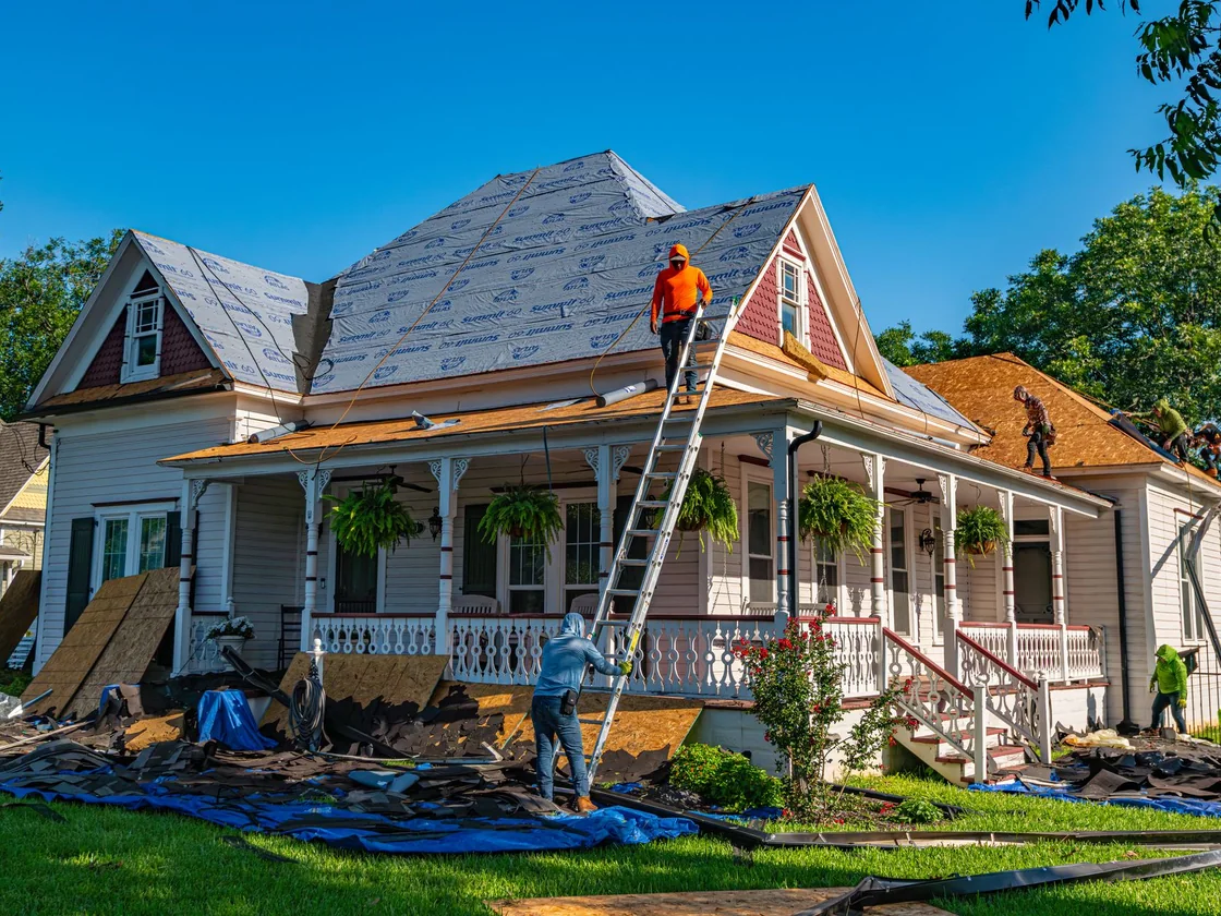 A roofing crew stripping shingles off a modest older frame house with torn-off shingles piled in the front yard