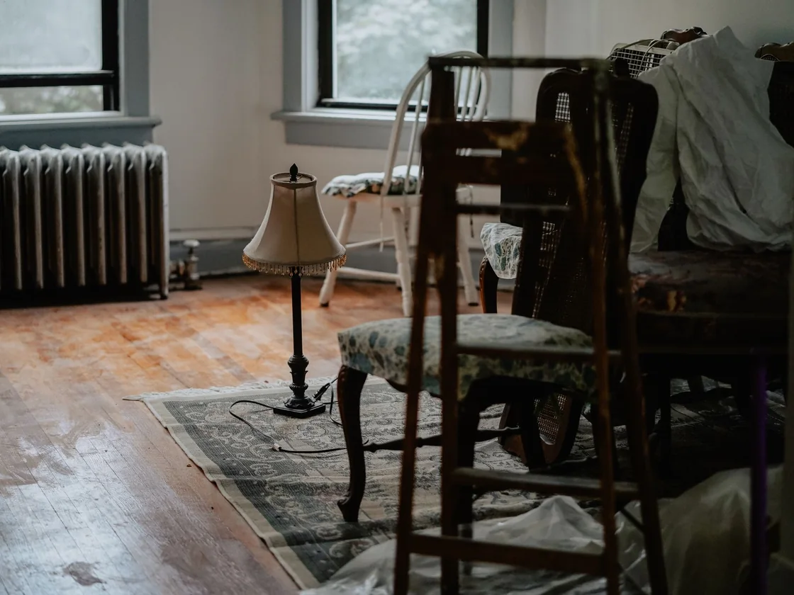 Old furniture, stacked chairs and a worn rug left behind in a dim room of an inherited house