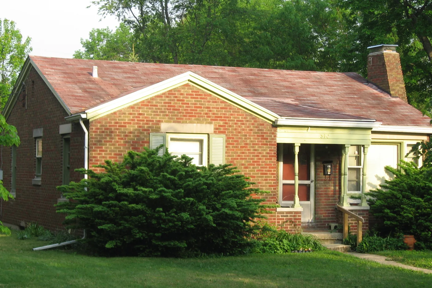 A small brick house with a worn roof, overgrown shrubs covering the front window and a homemade wooden handrail by the porch steps