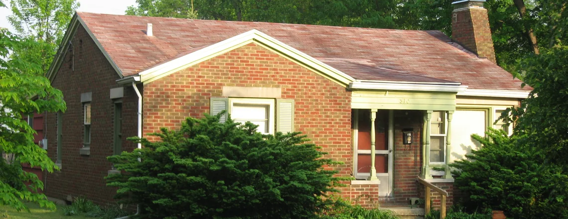 A small brick house with a worn roof, overgrown shrubs covering the front window and a homemade wooden handrail by the porch steps