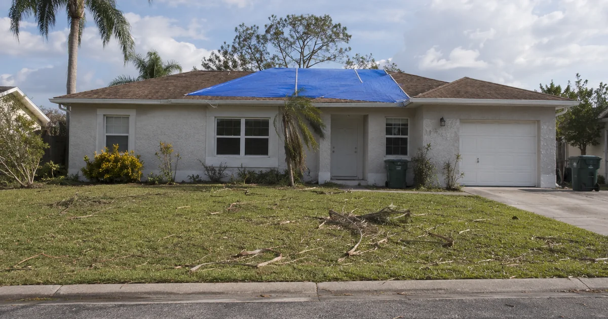 A modest single-family Florida house with a blue tarp on a storm-damaged roof under a clearing sky