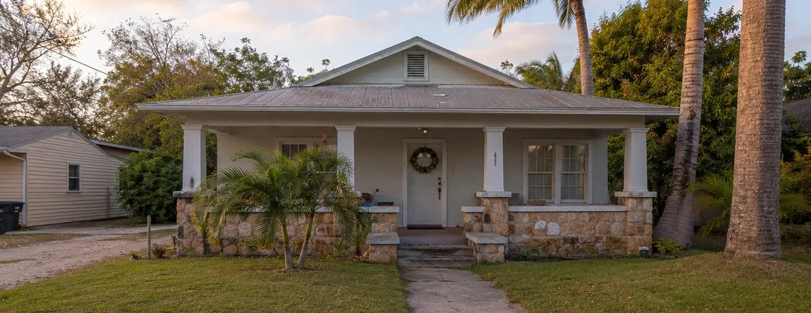 A modest older single-family Florida house with a wide front porch under palm trees, the kind of home often passed down to family