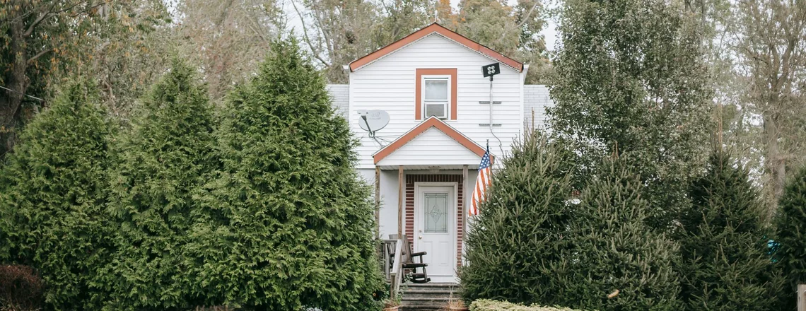 A modest white wood-frame house on an ordinary street with overgrown shrubs crowding the front porch