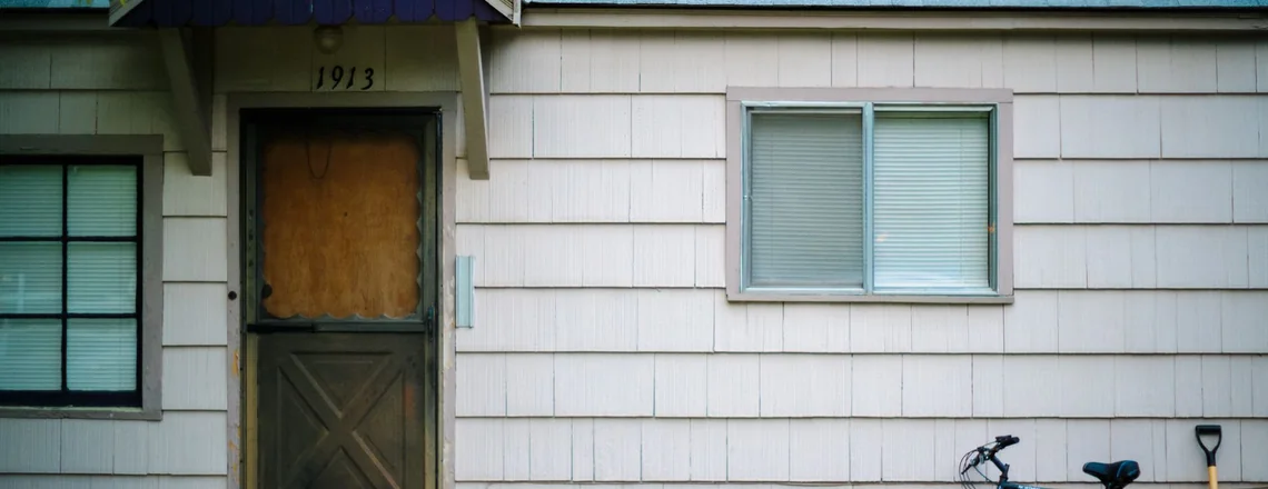 A small house with faded shingle siding, a worn wooden screen door and a bike leaning by the window on an ordinary street