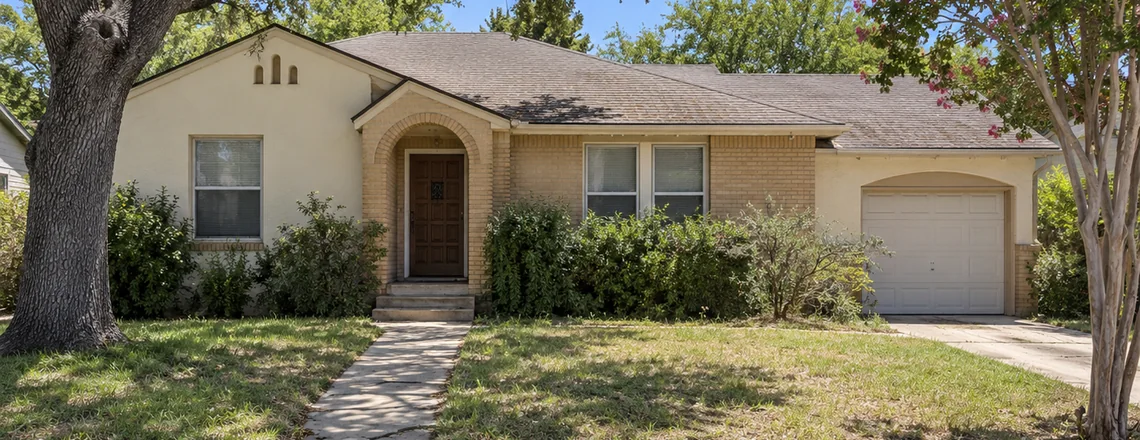 A modest one-story stucco and brick house with an arched entry and a tired front lawn on an ordinary San Antonio street