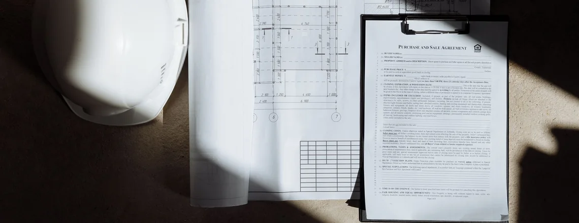 A hard hat, a set of building plans and a purchase and sale agreement on a clipboard laid out on a table
