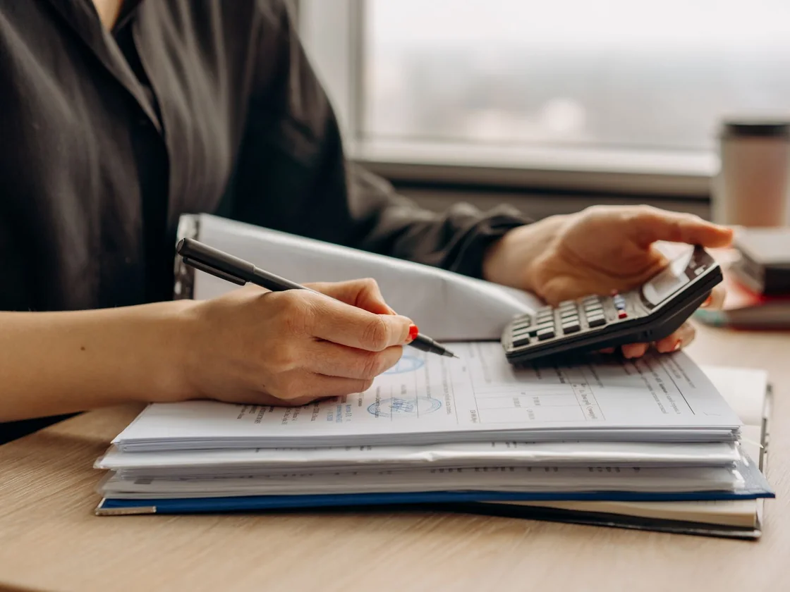 A person going through a stack of settlement paperwork with a red pen and a calculator