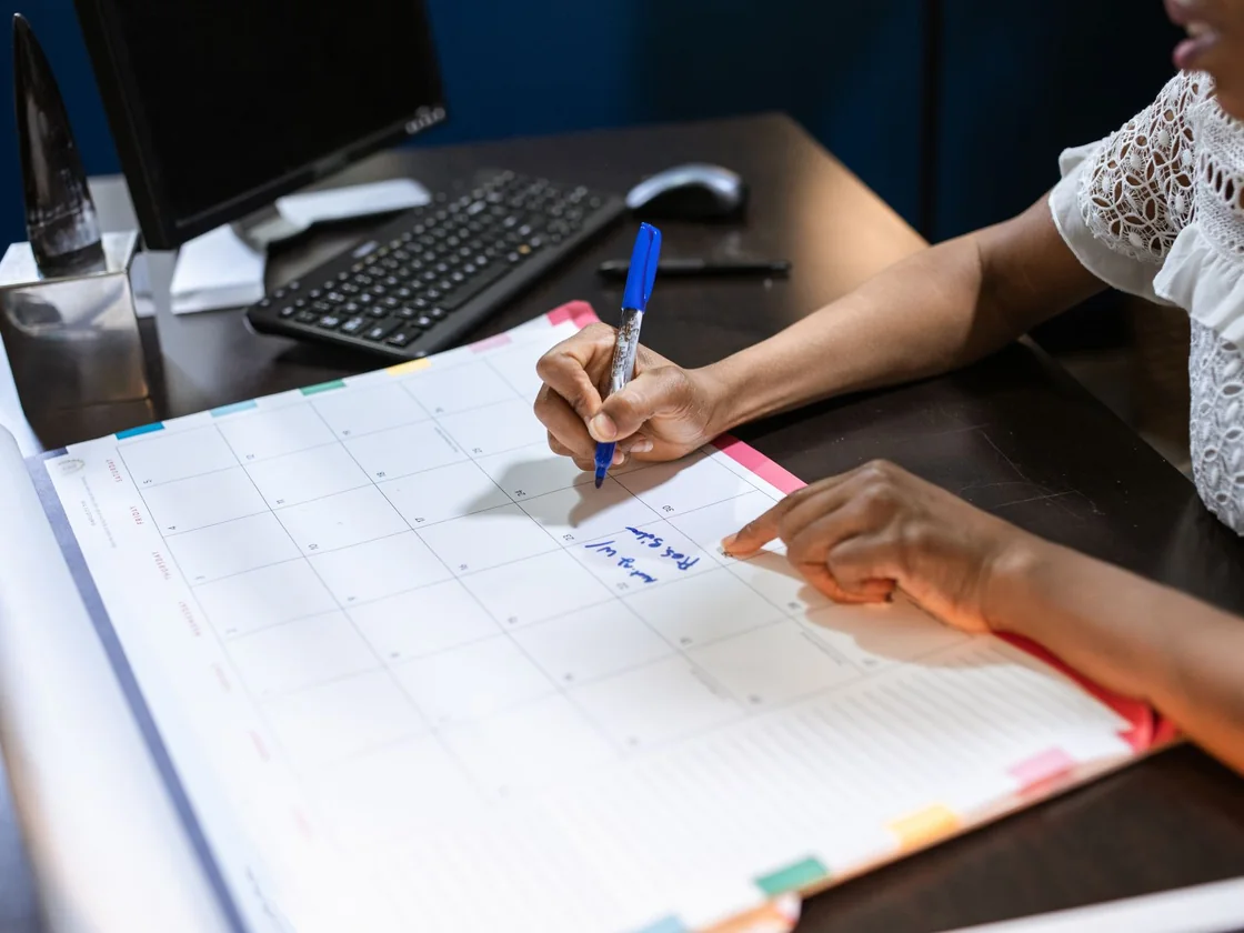 A hand marking a date on a desk calendar beside notes and a pen