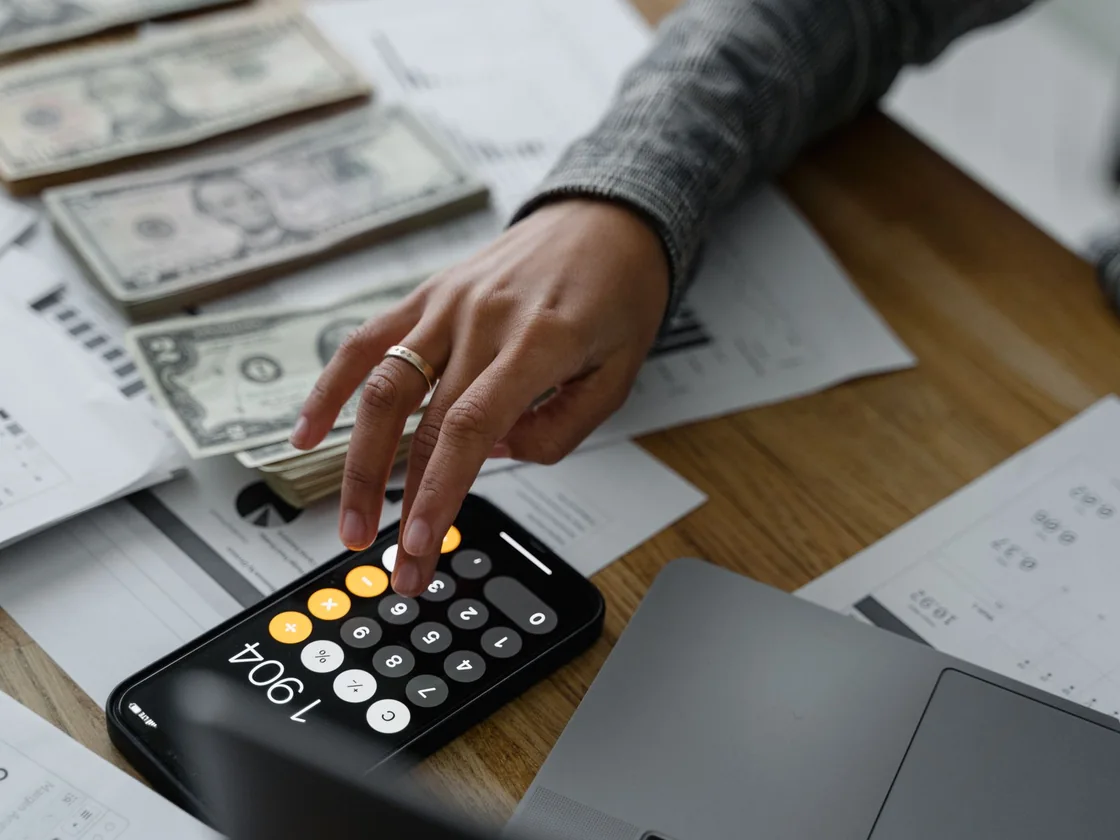 A hand working out sale proceeds on a calculator next to cash and paperwork on a table