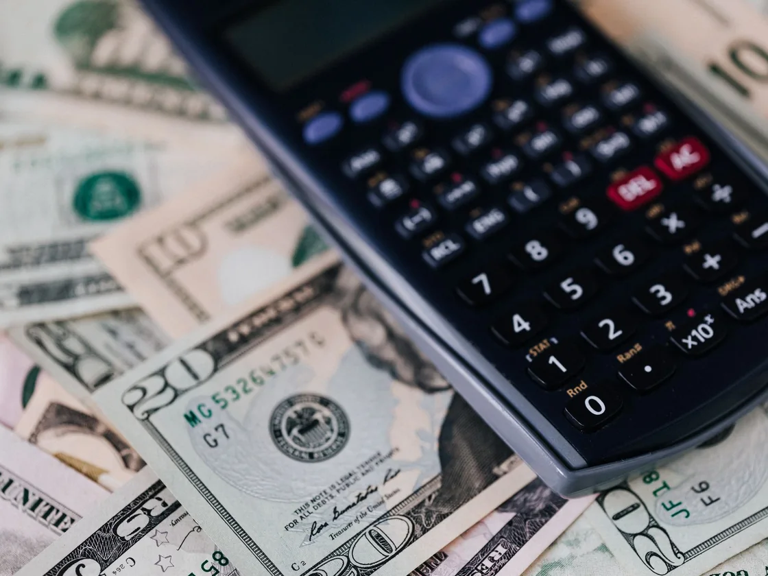 US dollar bills spread across a table with a calculator resting on top of them