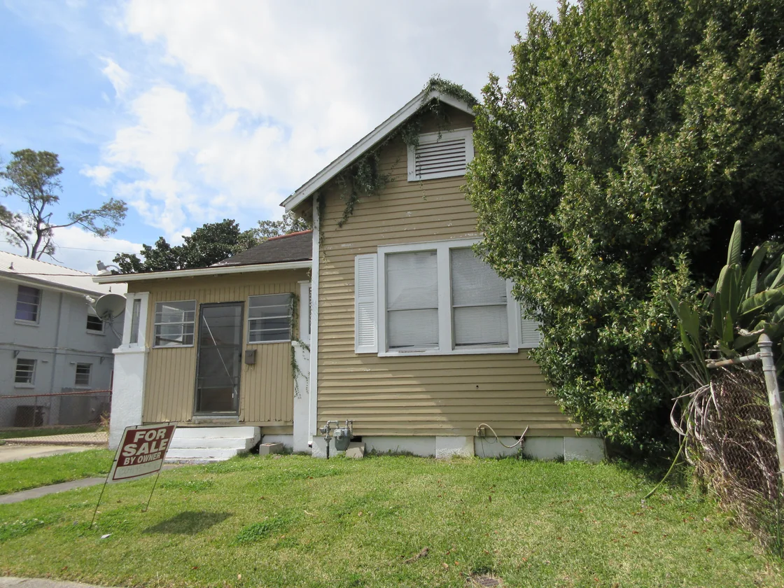A modest older house with a for sale by owner sign leaning in the front yard