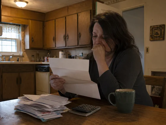 A homeowner at a kitchen table reading a foreclosure notice letter from the lender, with bills and a calculator nearby
