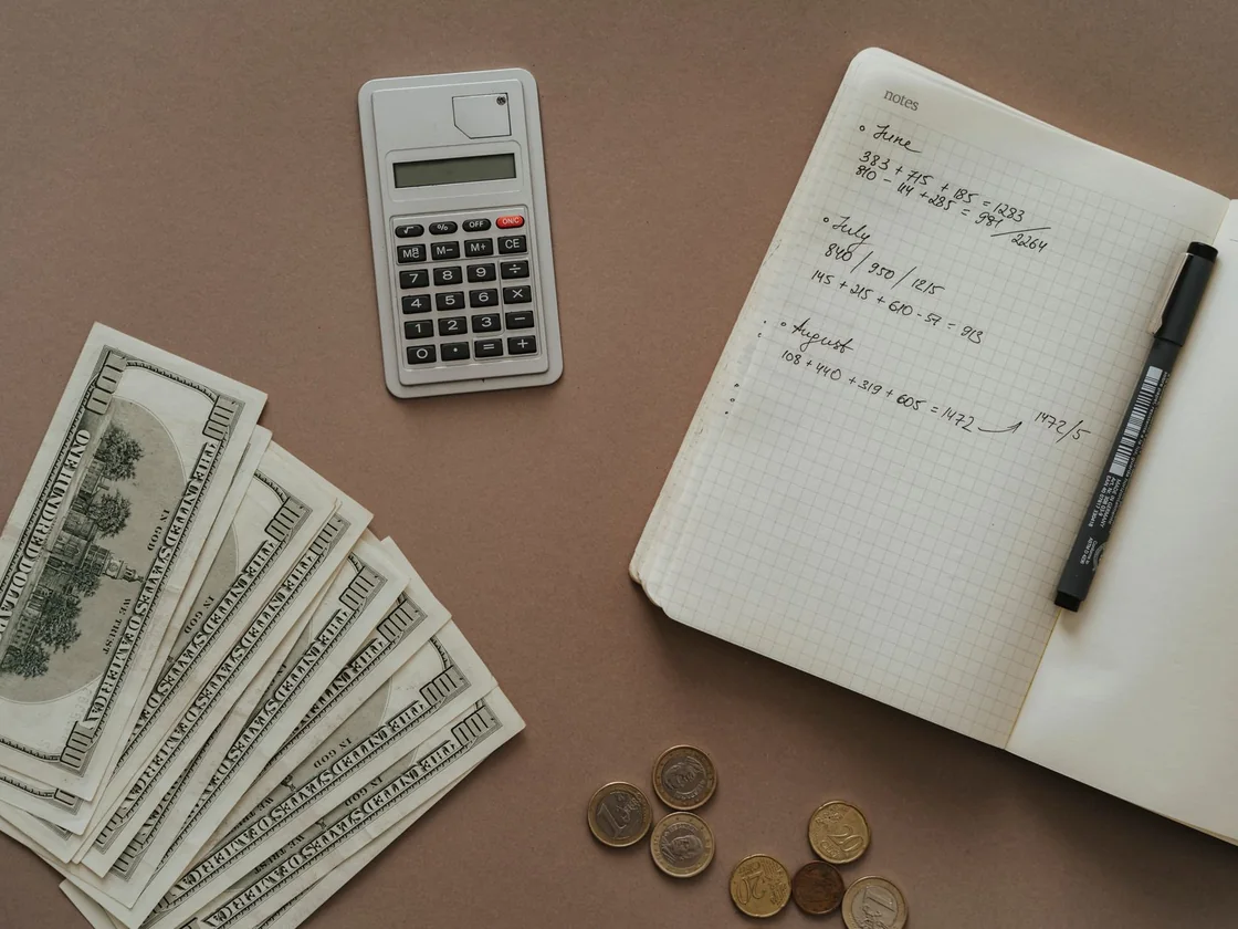 A calculator, a notebook and US dollar bills laid out on a table while someone works out the numbers