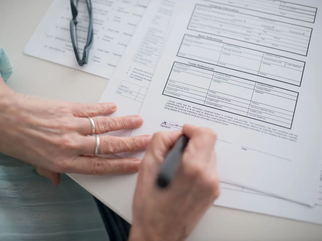 An older person&rsquo;s hands signing an official form at a table with reading glasses beside it