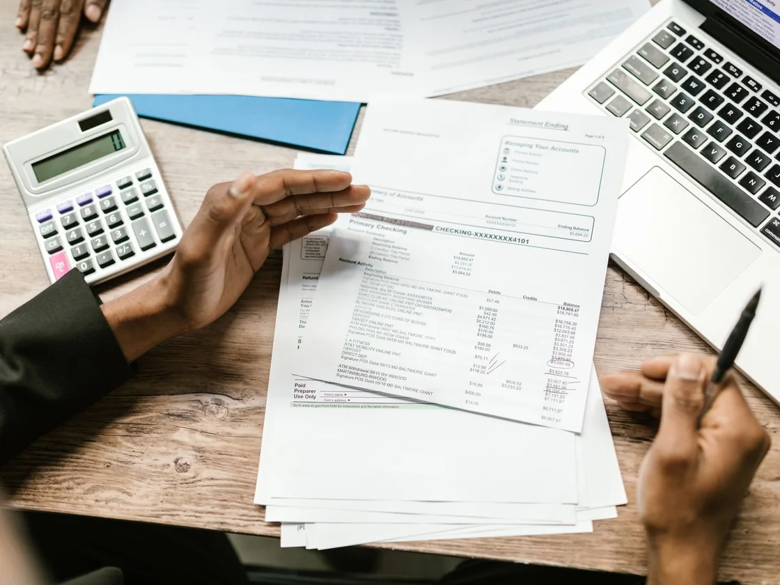 Hands holding a printed statement over a desk with a calculator, a pen and a laptop