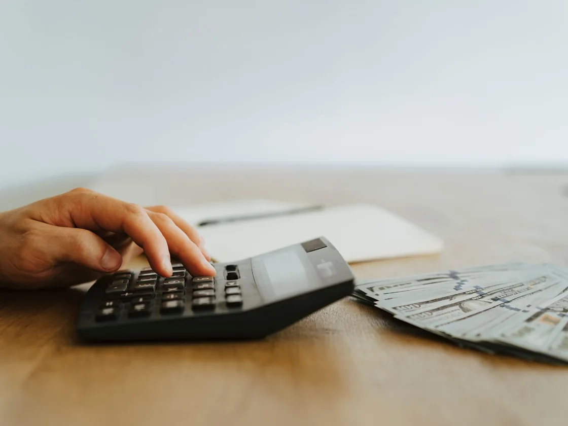 A hand working a desk calculator beside a fanned stack of US dollar bills and a notepad